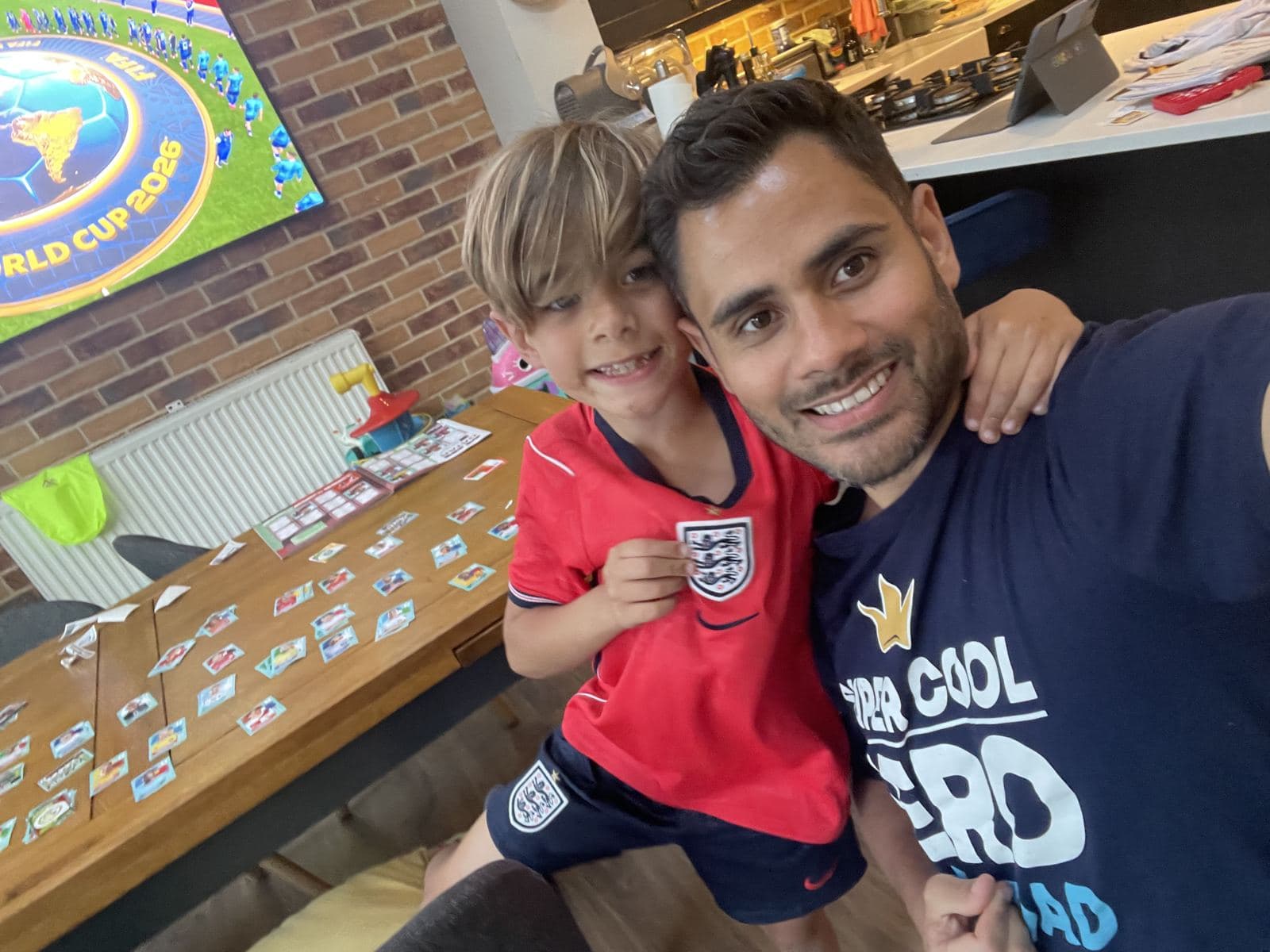 Jason and his son Jack smiling together at home, football stickers spread across the table and a match playing on the TV behind them